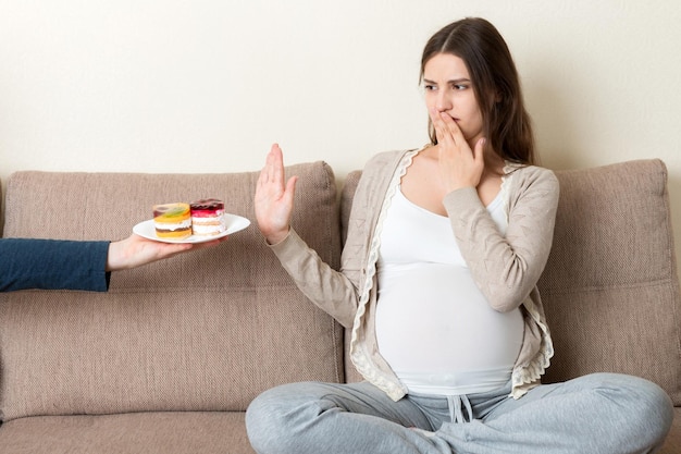Pregnant woman sitting on the sofa don't want to eat a cake and makes stop gesture No sweet and dessert during pregnancy concept