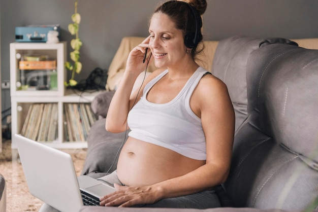 A pregnant woman sitting on the sofa with headphones and laptop working remotely