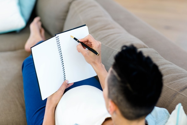 Pregnant woman sitting on sofa and writing on notepad