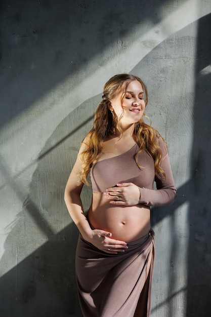 A pregnant woman in a skirt hugs her stomach with her hands Fashion pregnant woman Full length portrait on gray background Happy pregnancy