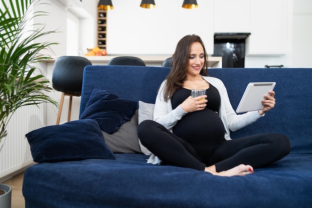 Беременная женщина в спортивной одежде сидит на синем диване в гостиной и пьет апельсиновый сок