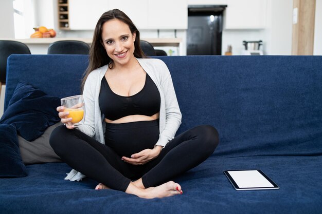 A pregnant woman in sports clothes sitting on a blue sofa in the living room and touching belly