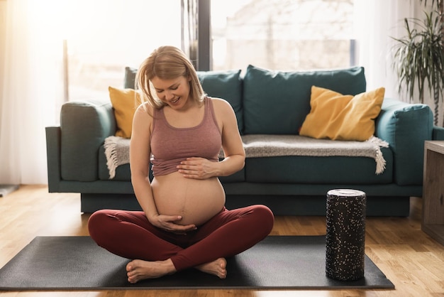 A pregnant woman in sports clothing is relaxing and exercising to promote wellbeing in her living room.