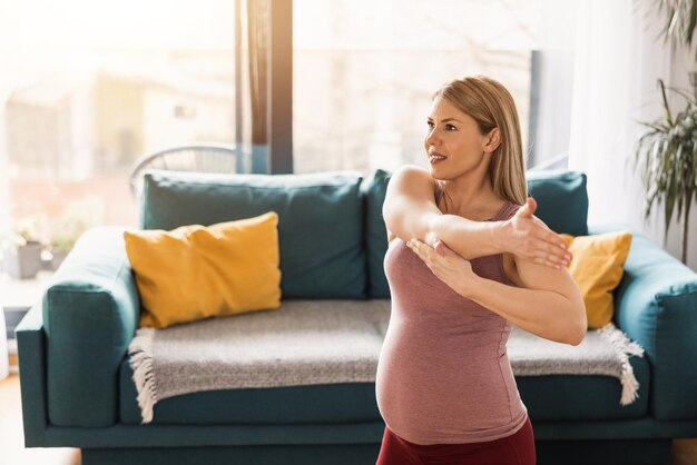 A pregnant woman in sports clothing is stretching and exercising to promote wellbeing in her living room.