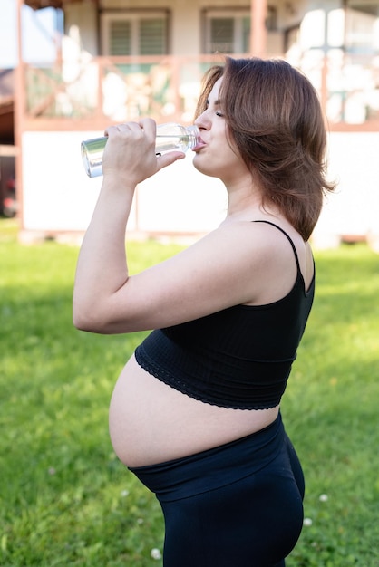 スポーツウェアを着た妊婦が外でヨガをした後,水のボトルを飲み,ヨガの練習に休憩し,フィットネスと瞑想をしています. 母性の精神健康と妊娠の概念