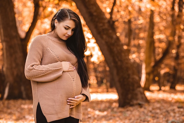 Pregnant woman standing in autumn city park forest, holding stroking her round belly with baby