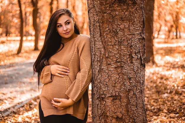 Pregnant woman standing in autumn city park forest, holding stroking her round belly with baby