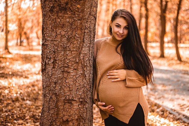 Pregnant woman standing in autumn city park forest, holding stroking her round belly with baby