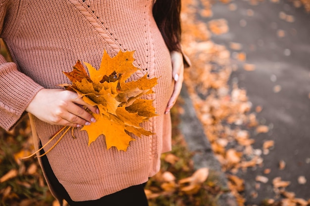 Pregnant woman standing in autumn city park forest, stroking her round belly with baby child inside