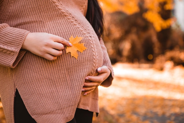 Pregnant woman standing in autumn city park forest, stroking her round belly with baby child inside