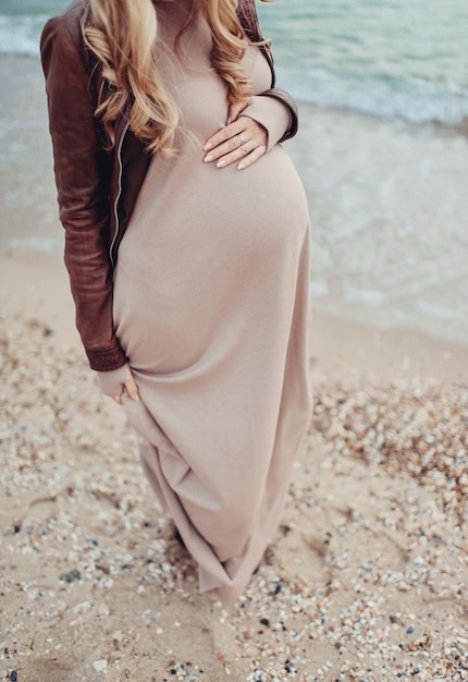 갈색 드레스와 갈색 가죽 재킷을 입고 해변에 서있는 임신한 여자