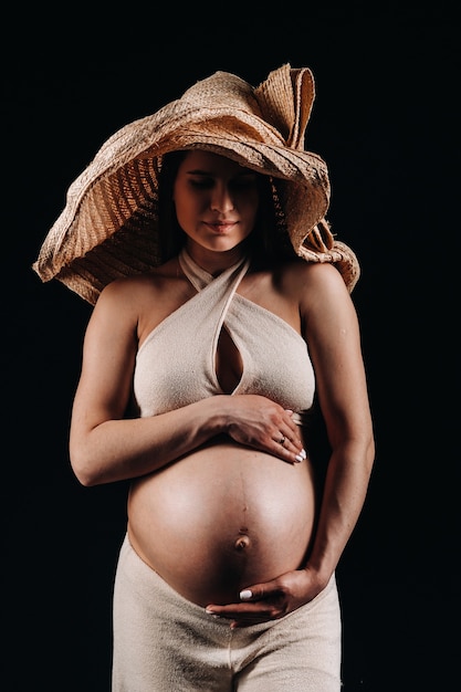 Pregnant woman in a straw hat in beige clothes in the studio on a black background.