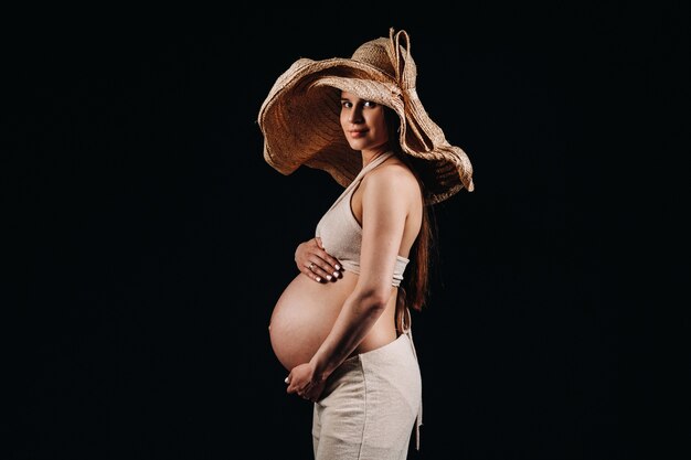 Pregnant woman in a straw hat in beige clothes in the studio on a black background.