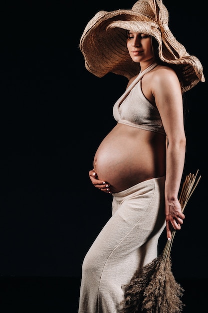 黒の背景のスタジオで彼女の手に花束とベージュの服を着た麦わら帽子の妊娠中の女性