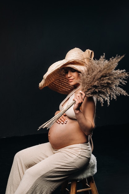 Pregnant woman in a straw hat with beige clothes with a bouquet in her hands in the studio on a black background.