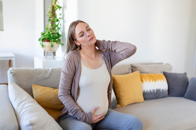 Pregnant woman suffer nrck and shoulder pain. pregnant woman holding her injured neck while touching her belly and sitting on couch