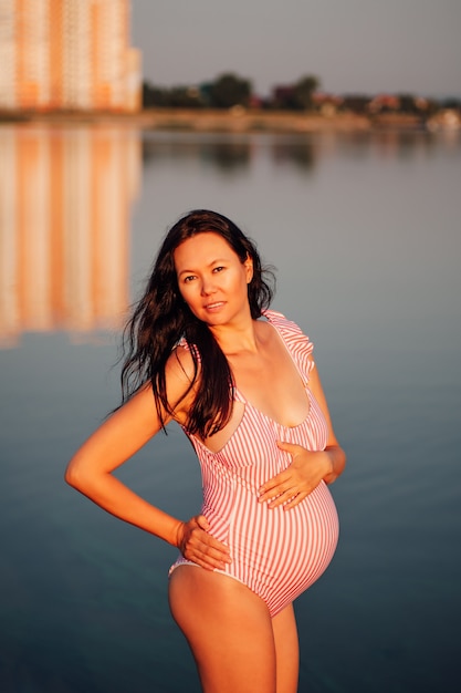 Pregnant woman in a swimsuit relaxed darkhaired pregnant woman in a pink swimsuit holds her hands on...