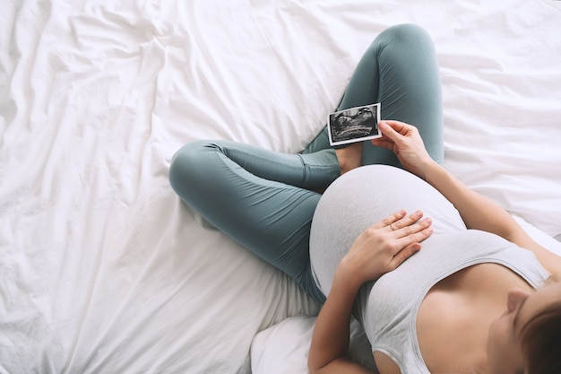 Pregnant woman touching her belly looking at ultrasound image in bed