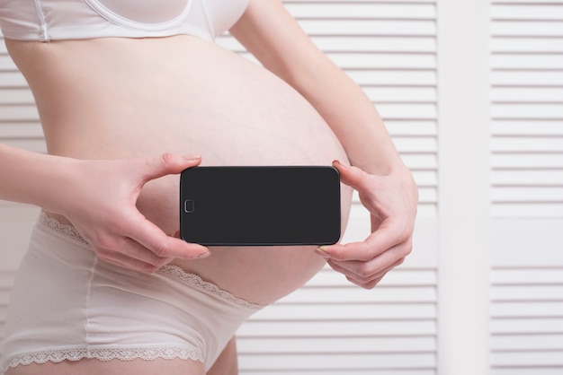 Pregnant woman in underwear holding and showing mobile phone with empty display