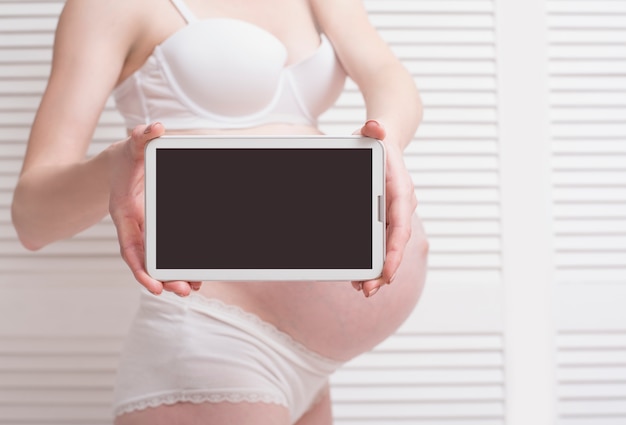 Pregnant woman in underwear showing digital tablet with empty display