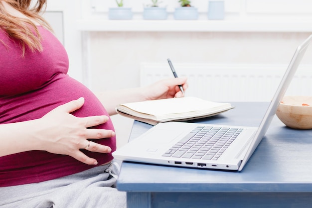 A pregnant woman uses a laptop and makes notes in a notebook The concept of working at home