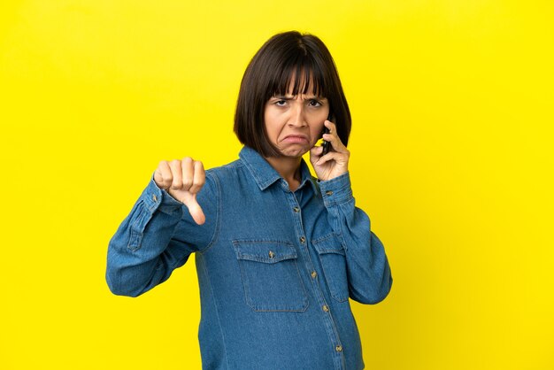 사진 부정적인 표현으로 아래로 엄지 손가락을 보여주는 노란색 배경에 고립 된 휴대 전화를 사용하는 임신 한 여자