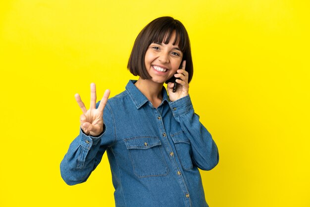 Donna incinta che utilizza il telefono cellulare isolato su sfondo giallo felice e conta tre con le dita