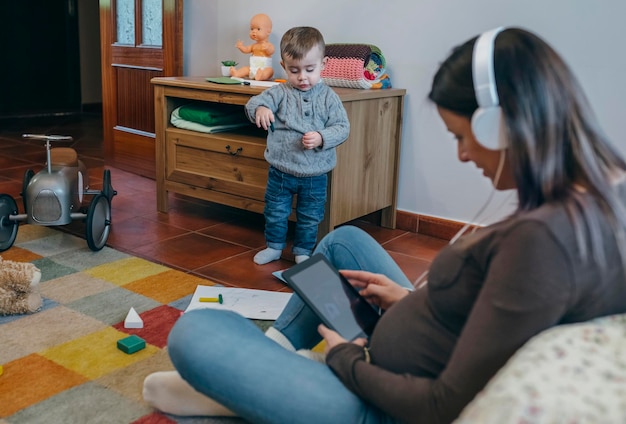 息子が遊んでいる間、リビングルームのカーペットの上に座っているタブレットを使用している妊婦