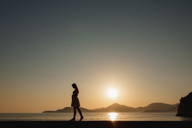 A pregnant woman walks on the seashore at sunset and gently strokes her belly, behind her are mountains and the setting sun . 