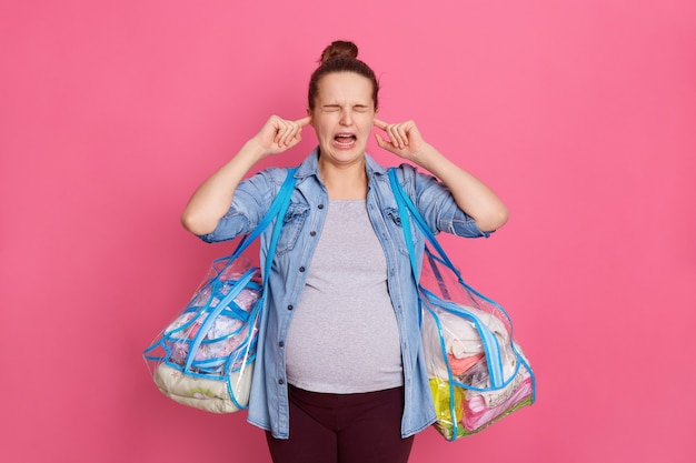 Pregnant woman wearing casual clothing covering both ears with fingers isolated on pink