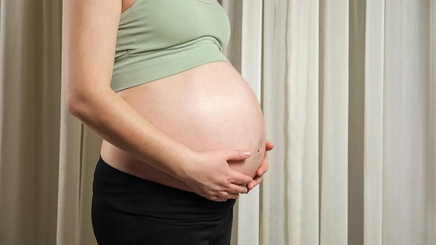 Pregnant woman wearing sports outfit touches stroking bare belly caressing with loving hands standing against window curtain closeup.