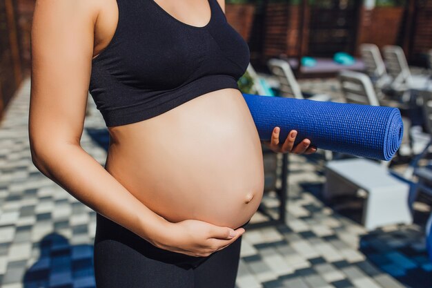 Una donna incinta che indossa abbigliamento sportivo e un tappetino da yoga all'aperto le tiene la pancia e va allo yoga