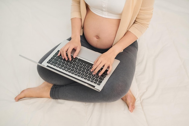 Беременная женщина в белой одежде сидит и использует ноутбук в постели
