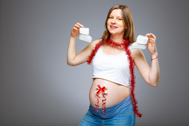 아기 옷과 크리스마스 이브에 아기 birt를 기다리는 그녀의 배꼽에 빨간 활을 가진 임신한 여자