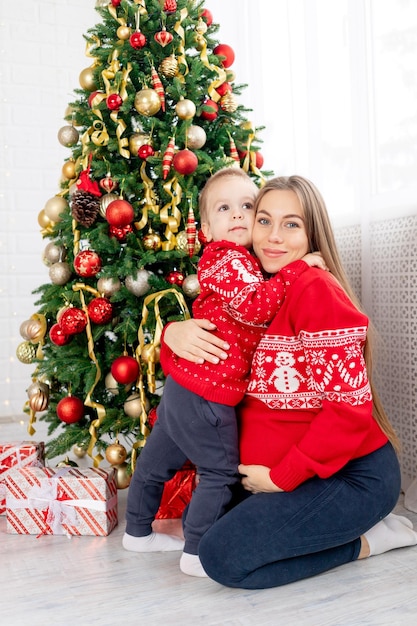 A pregnant woman with a baby boy in a red sweater under the Christmas tree at home hugging congratulating each other and enjoying the new year and Christmas