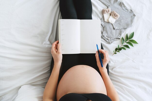 Photo pregnant woman with beautiful belly makes notes or check list in paper diary concepts of preparation for baby birth tips for a healthy pregnancy minimalist style photography closeup indoors