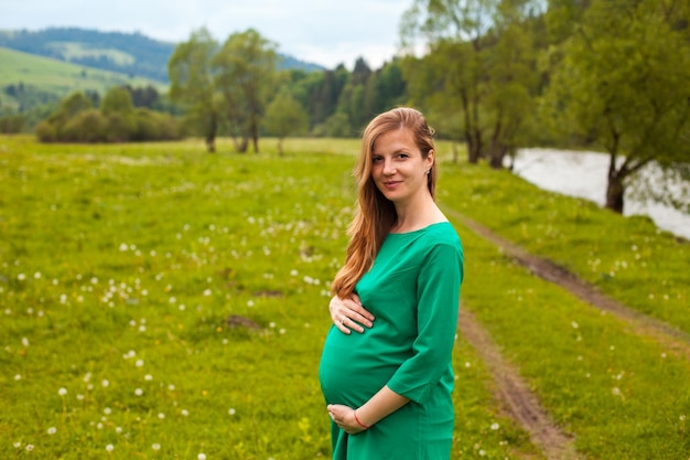 緑の美しいチュニックで美しい茶色の髪の妊婦は、緑の木々と自然の背景に澄んだ空気を呼吸しています