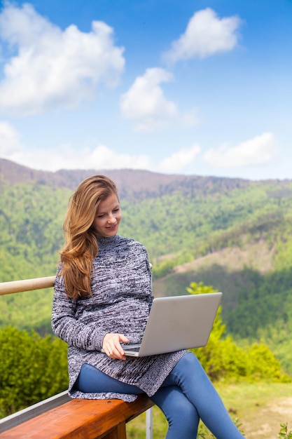 La donna incinta con bei capelli e con il computer portatile è seduta sullo sfondo della natura pacifica