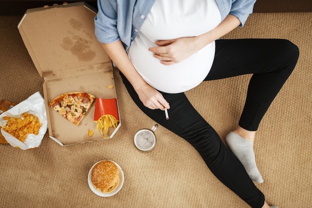 腹を吸って、家で不健康な食べ物を食べる妊婦