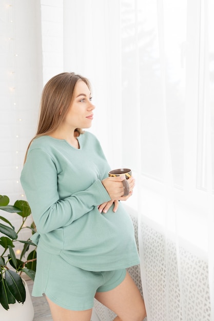 A pregnant woman with a big belly strokes him standing at the window in the morning with a mug of tea, the concept of waiting for a child, love and upbringing