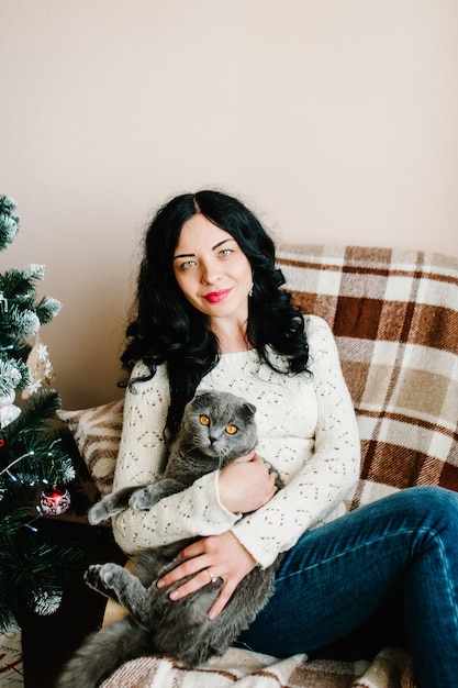 Pregnant woman with cat posing near christmas tree at home.\
merry christmas and happy holidays! pregnancy, holidays, people and\
expectation concept.