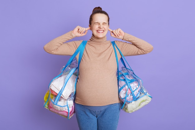 Pregnant woman with hair bun wearing beige sweater and jeans, covering both ears with hands