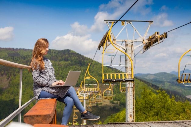 Pregnant woman with long hair and with laptop is sitting on the background of ski lift