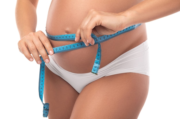 Pregnant woman with measuring tape is checking her belly girth isolated on white wall