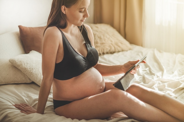 Беременная женщина с современным планшетом сидит на кровати и поправляет длинные волосы