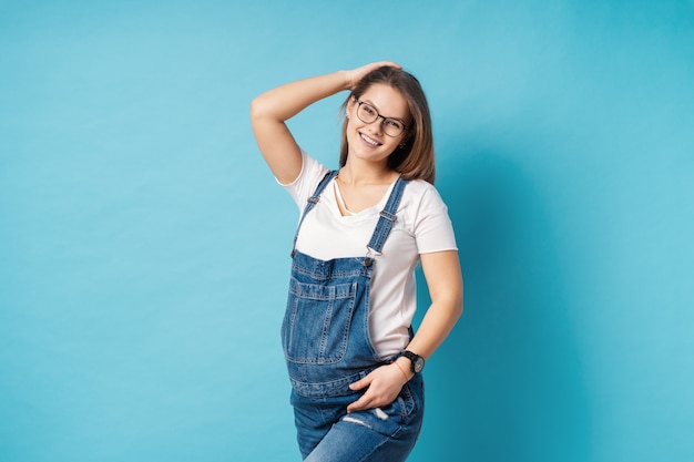 Pregnant woman, with stylish glasses, touching her hair, smiling isolated on blue background in studio