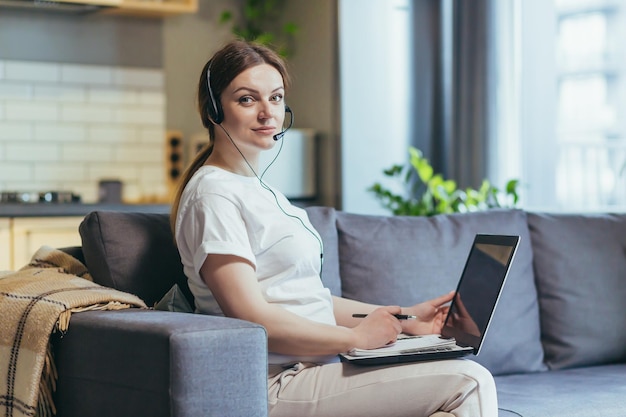 Pregnant woman working remotely at home using video call headset consultant sitting on sofa and working with laptop at home