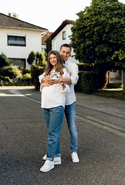 a pregnant woman and a young husband are hugging while holding an ultrasound scan near the pregnant