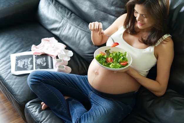 Photo pregnant women sitting on sofa is holding salad bowl in her hand. her future born baby dress