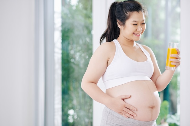 妊娠中の若いアジアの女性は、新鮮なオレンジジュースを飲み、朝の運動後に腹に触れます
